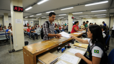 Homem brasileiro entregando documentos de transferência veicular para atendente no balcão do DETRAN com fila de espera ao fundo